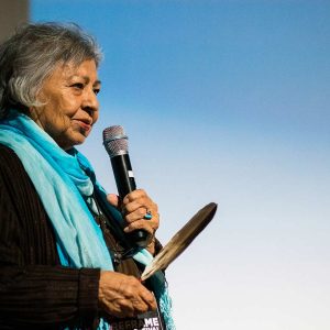 Elder Shirley Williams speaking into a microphone on stage, holding an eagle feather