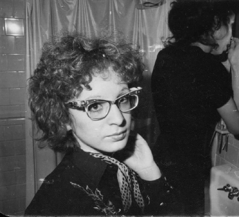 A black and white image of Nan Goldin who stares into the camera. They have short curly hair and glasses and are wearing a polka dot scarf and collared shirt. Their left hand is raised to their cheek. In the background, a person gets ready in a bathroom mirror.