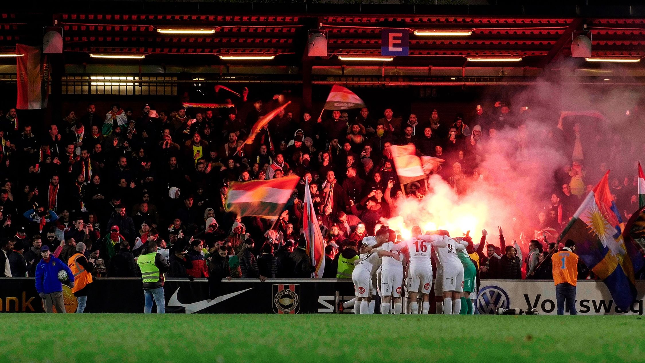 A group of soccer players are huddled together, celebrating near the edge of the field. They are surrounded by vibrant flares and smoke, mostly in red, adding to the dramatic atmosphere. In the background, the crowd is animated, with some fans waving flags energetically. The scene is at night, under artificial lighting, and captures the energy and excitement typical of a soccer match.