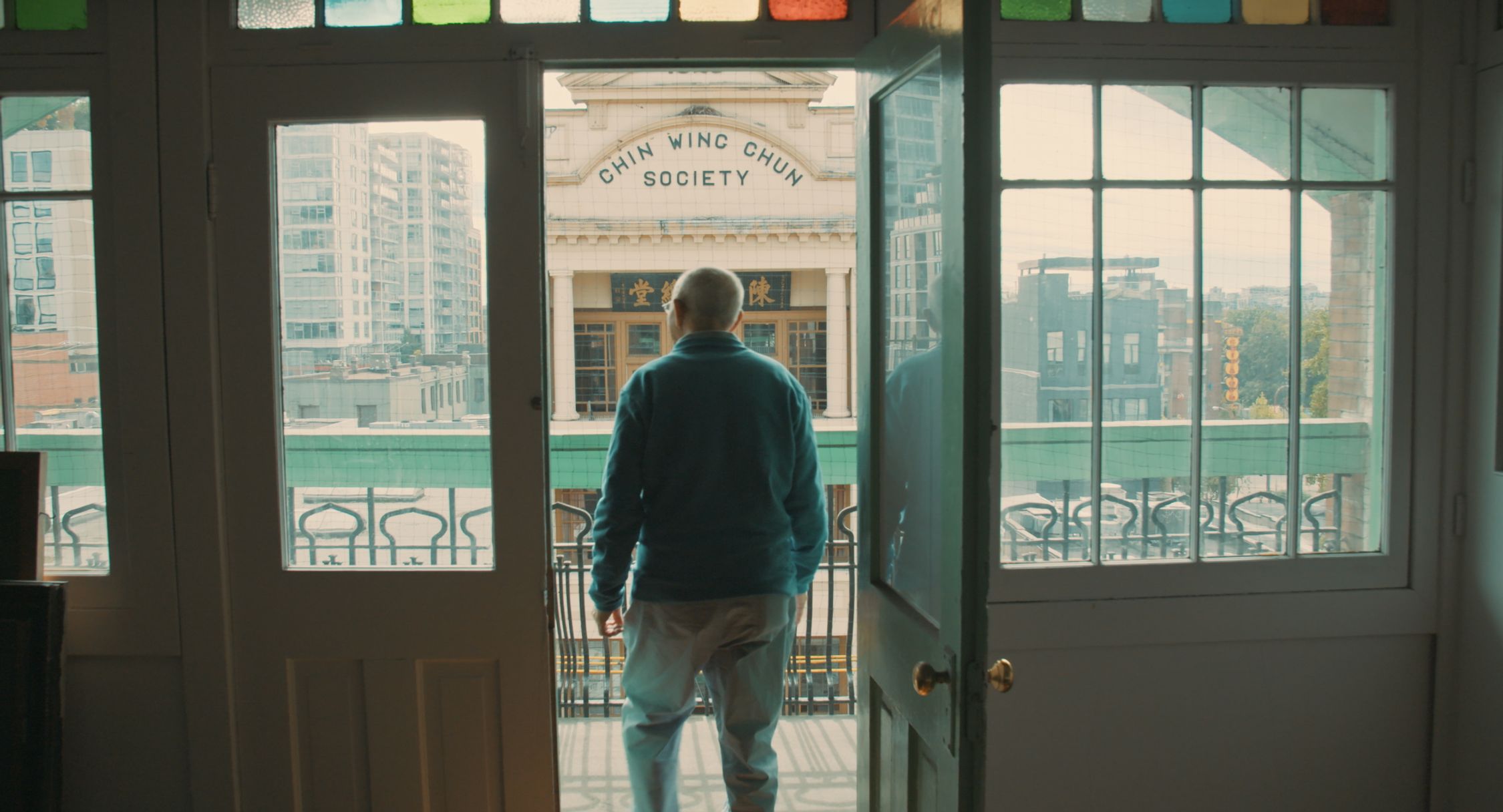 An individual is standing at an open doorway, facing a balcony and looking out at an urban landscape. The sign above the balcony reads "CHIN WING CHUN SOCIETY" in English and has Chinese characters beneath it. The view includes modern buildings, and the doorway is framed with colourful glass panes, suggesting a traditional or historical setting within a contemporary city environment.