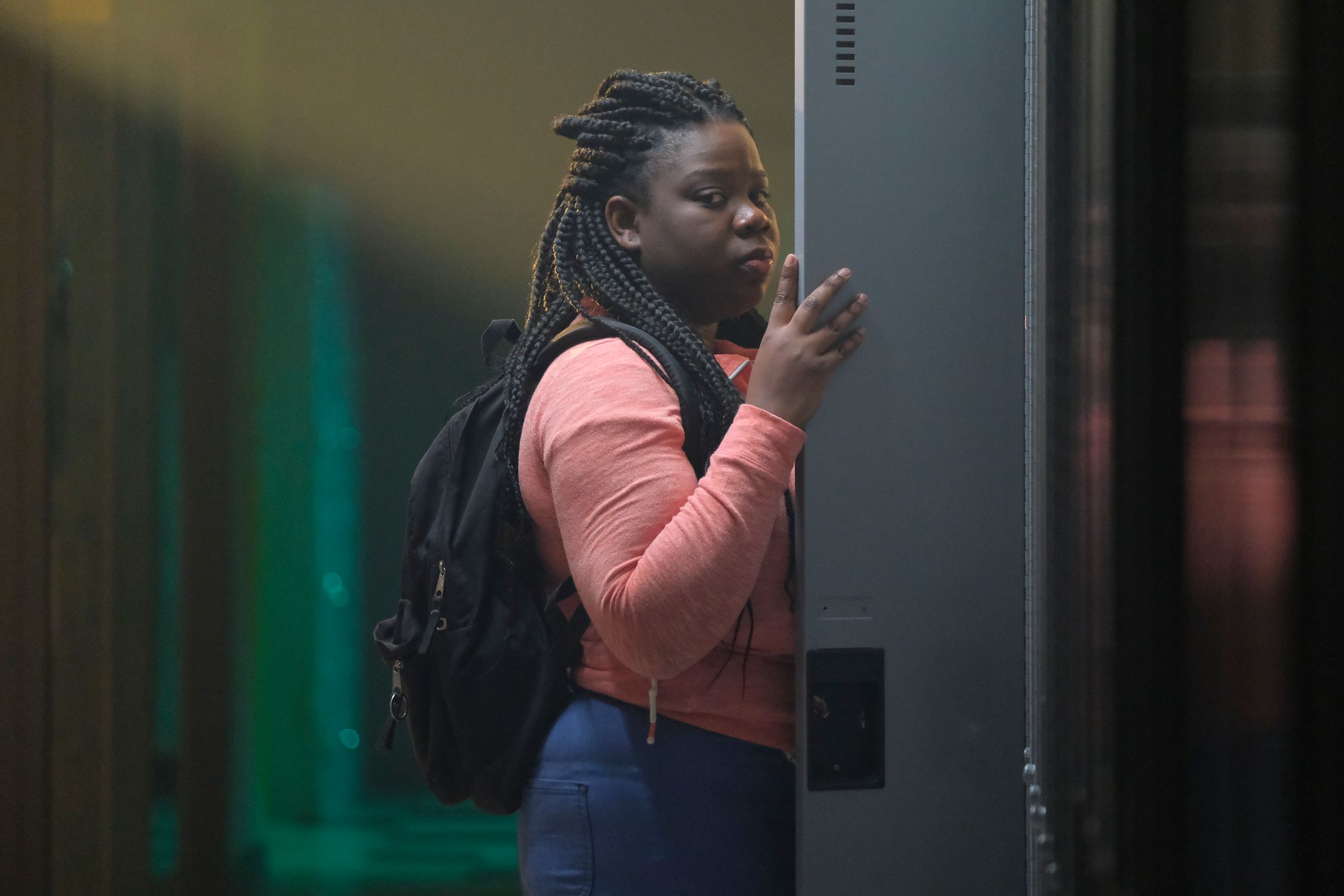 A person with braided hair is standing at a locker, looking over their shoulder with a contemplative expression. They are wearing casual clothing and a backpack, indicating they could be a student. The hallway in the background is blurred.
