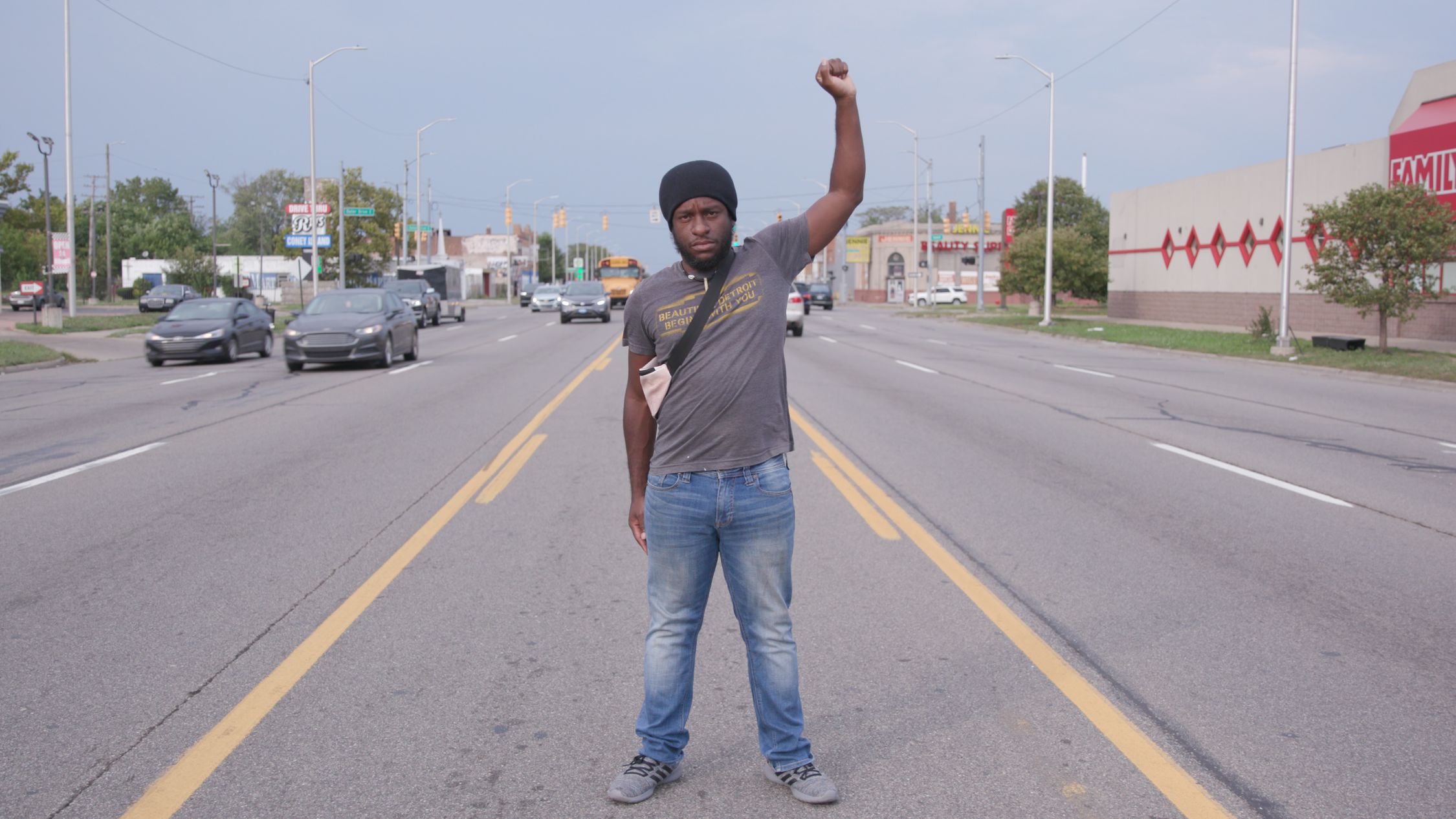 A person is standing in the center of a street with one fist raised high. The individual is wearing a dark beanie, a t-shirt, and jeans, and has a serious expression. The street is lined with various commercial buildings and cars are visible in both directions. The context of the raised fist, a symbol often associated with solidarity and support for a cause, suggests a moment of protest or statement. The image captures a sense of defiance or activism.