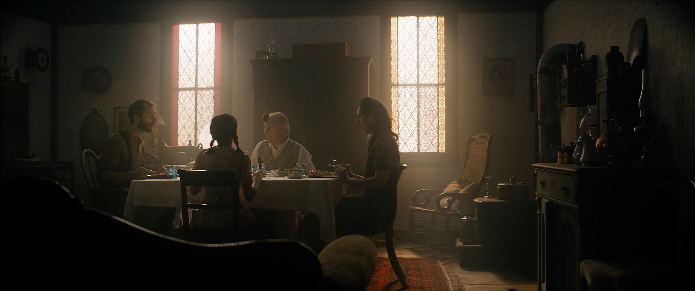 Four individuals are seated around a dining table in a dimly lit room that suggests a vintage or historical setting. The warm glow from the windows, adorned with patterned stained glass, provides natural light. The room is decorated with patterned wallpaper, and there are traditional furnishings and decorations, including a cabinet and a clock on the wall, which contribute to the room's old-fashioned ambiance. The scene has a calm and intimate feel, with the focus on the people at the table, possibly engaged in conversation over a meal.
