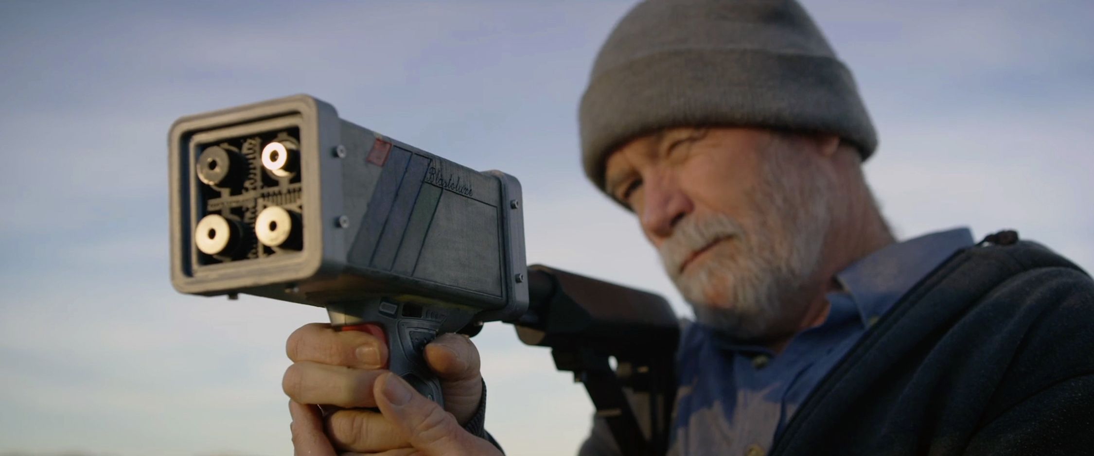 An individual, likely a mature adult given the visible gray beard and wrinkles, is aiming a laser gun. The device is held up to their face, and their focused gaze is directed towards the target. They are wearing a gray beanie and a dark jacket, and the background is softly blurred with a clear sky, indicating an outdoor setting during the day. The device’s detailed mechanics are visible, emphasizing a the advanced technology.
