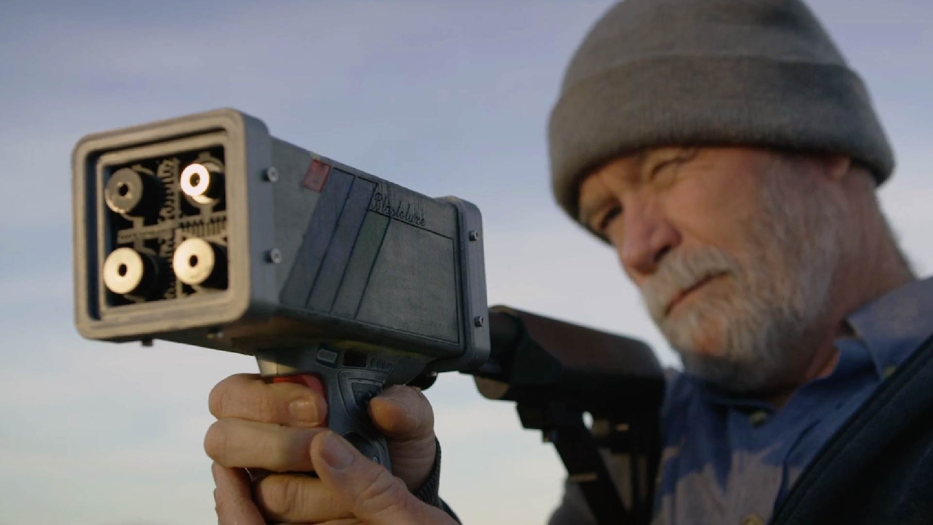 An individual, likely a mature adult given the visible gray beard and wrinkles, is aiming a high-tech drone suppression device. The device is held up to their face, and their focused gaze is directed towards the target. They are wearing a gray beanie and a dark jacket, and the background is softly blurred with a clear sky, indicating an outdoor setting during the day. The device's detailed mechanics are visible, emphasizing a the advanced technology.