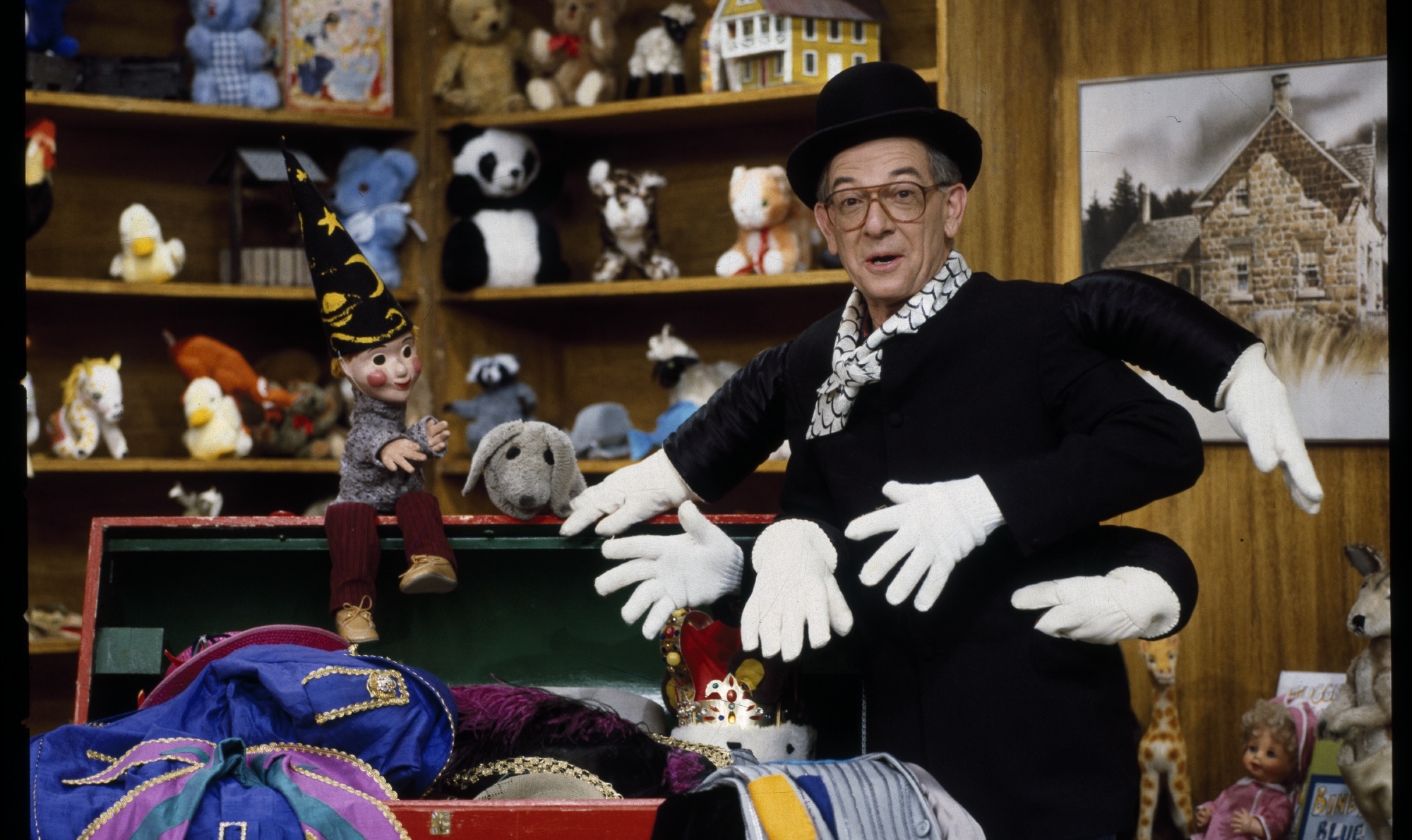 An image of a person wearing a top hat, glasses, and black and white scarf. To their left is a red tickle trunk with flowers painted onto it. The trunk is full of colourful clothes spilling over the edge. The person has on a fantastical black top, making it seem like they have six arms with white gloves covering the hands. In the background are shelves full of stuffed animals and other toys.