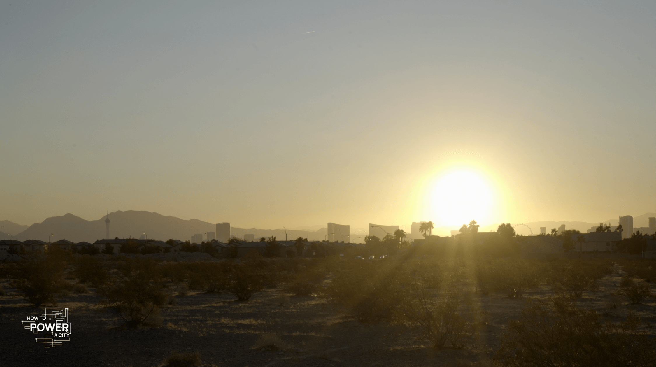 A sunset over a desert cityscape with the sun low in the sky, casting a warm glow and creating a silhouette of the mountains in the background. There are outlines of buildings and palm trees against the light, and the foreground shows arid terrain with sparse vegetation typical of a desert. In the lower left corner, there's text that reads "HOW TO POWER A CITY," suggesting the image may be related to a documentary or educational content focusing on urban energy topics. The sky above is clear with minimal clouds, allowing the sunlight to dominate the scene.