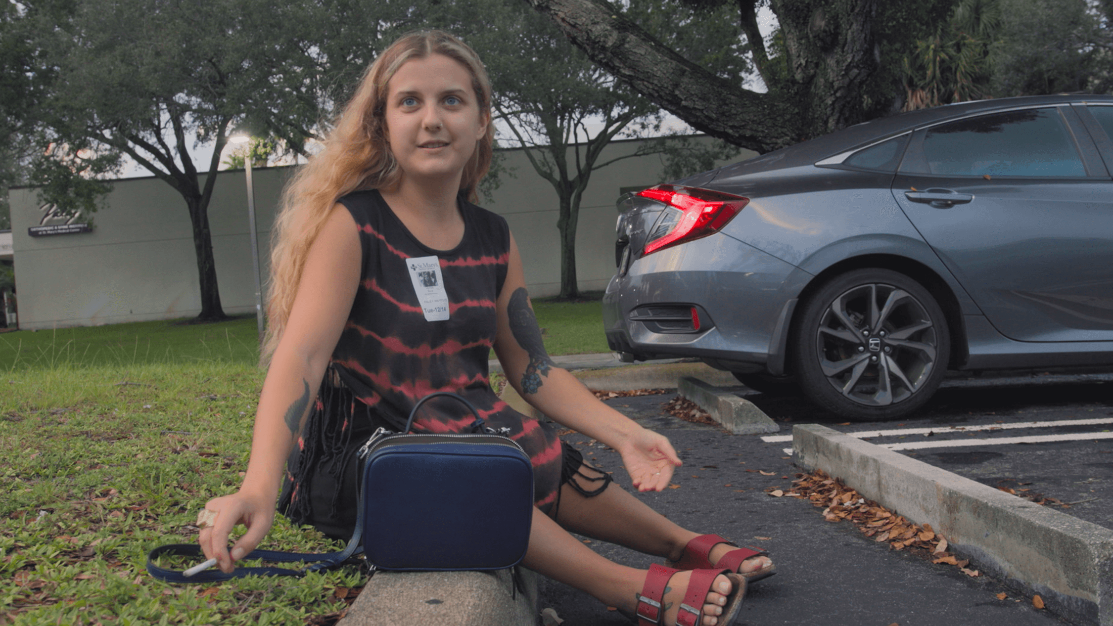 A person is seated on a curb in a parking lot, wearing a tie-dye dress and sandals, with a small blue handbag next to them. They have a tattoo on their arm and are wearing a name tag sticker. The person looks off to the side with a thoughtful expression. Behind them, there is a car parked and trees that suggest the area could be near an office or a public building. The setting appears to be during the day with overcast weather.