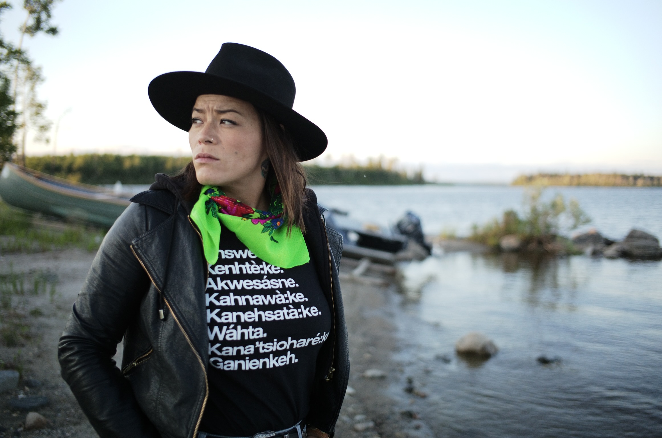 A person with a solemn expression stands by a lakeshore, wearing a black hat, a black T-shirt with white text, and a bright green scarf with red accents. The T-shirt's text includes Indigenous community names. In the background, there's a blurred view of a canoe on the shore and a motorboat in the water. The setting suggests a serene, natural environment during the daytime.