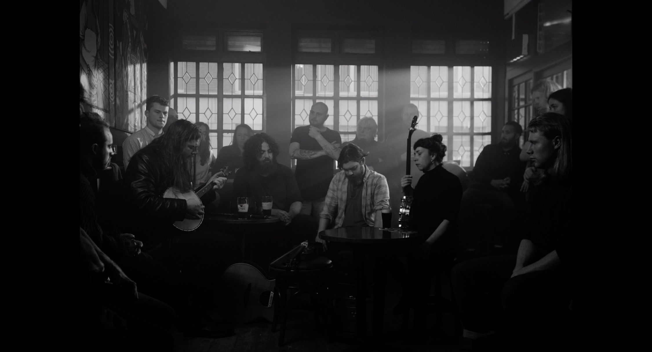 In a dimly lit pub, a group of people are gathered, some sitting and some standing. The focus is on a person playing a banjo in the center, surrounded by onlookers who are attentively listening or waiting their turn to perform. The mood is casual and communal, typical of an informal music session. The image is monochromatic, which adds a timeless and candid feel to the scene.
