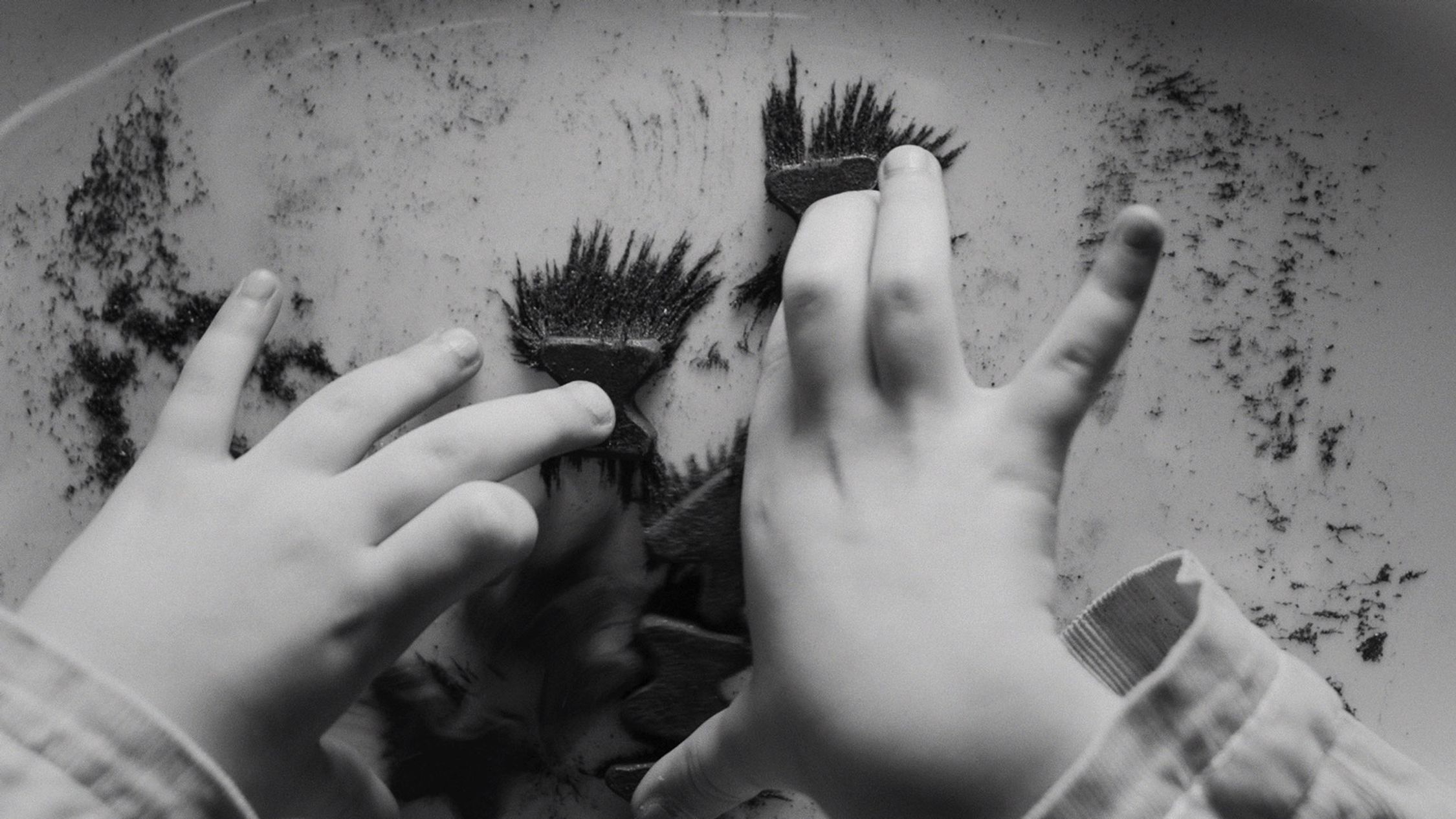 The image is a monochrome photograph of a child's hands interacting with magnets. The hands are small with youthful features, suggesting the person's young age. The magnets on the fingertips have attracted iron filings, which have clustered around the poles of the magnets, creating a visual display of the magnetic field lines. The activity seems to be an exploratory or educational play with the principles of magnetism.