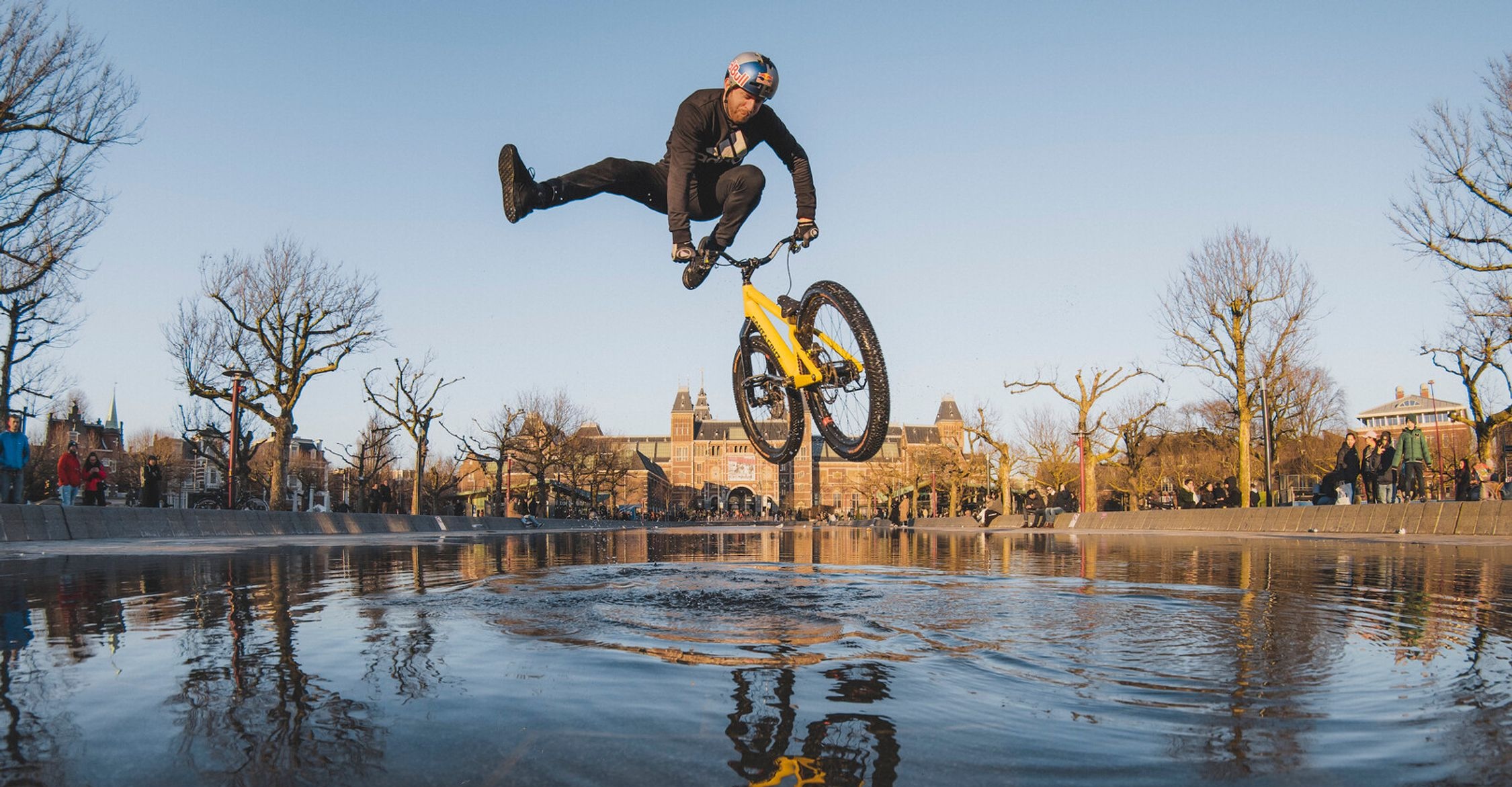 A BMX rider is captured mid-air performing a stunt, with their body horizontal to the ground and legs kicked out to the side, above a yellow bicycle. The bike's reflection is visible on the wet surface below. The shot is set against an urban park backdrop with historical buildings, bare trees, and a clear blue sky. Onlookers are seen in the distance. The low angle of the photograph emphasizes the height of the jump and the skill of the rider.