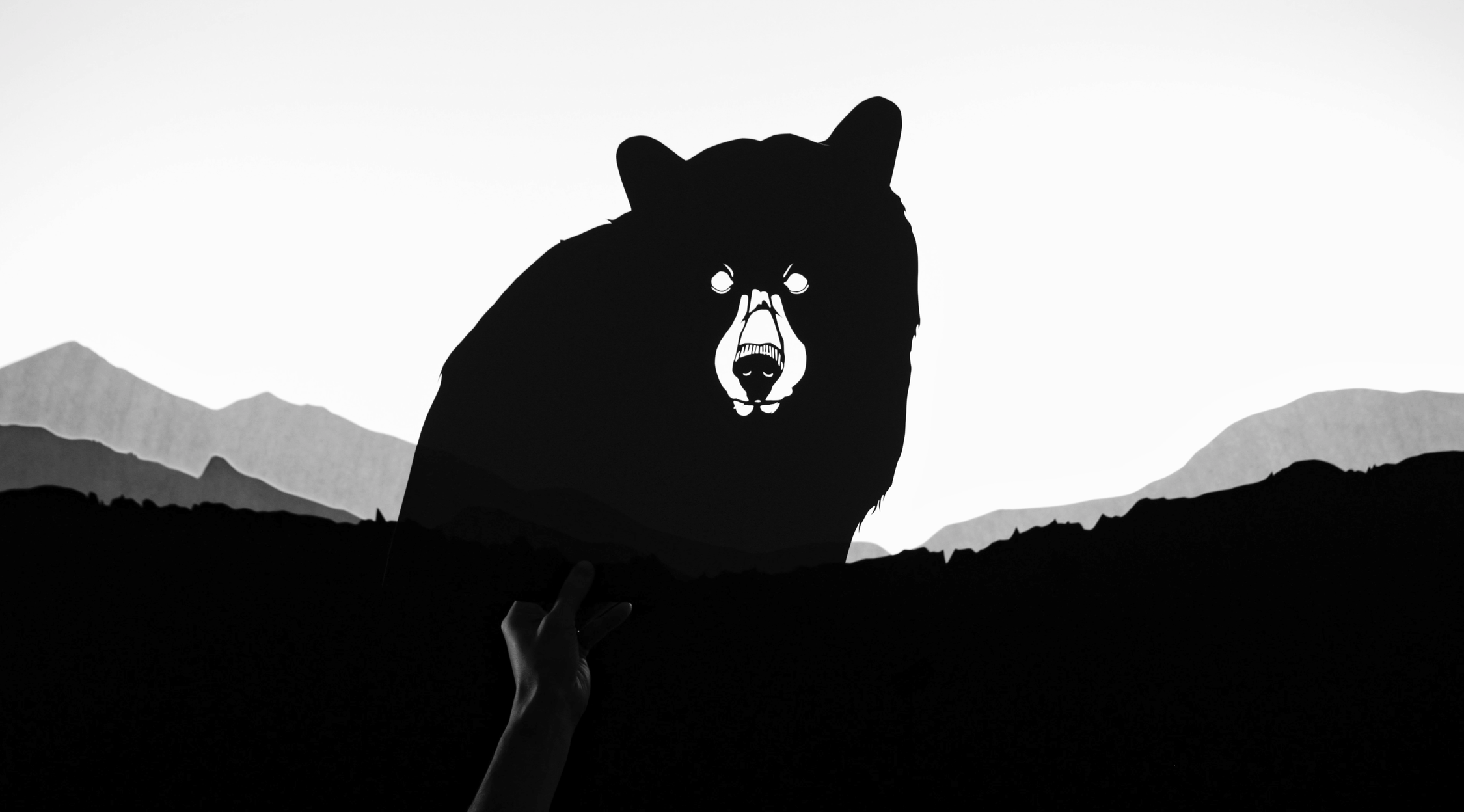 Silhouetted against a light sky with faint mountain outlines, there is a large, stylized black cutout of a bear with white accents for the bear's eyes, nose, and mouth, giving it a stark, graphic appearance. In the lower left corner, a human hand is visible, holding the bottom edge of the bear silhouette.