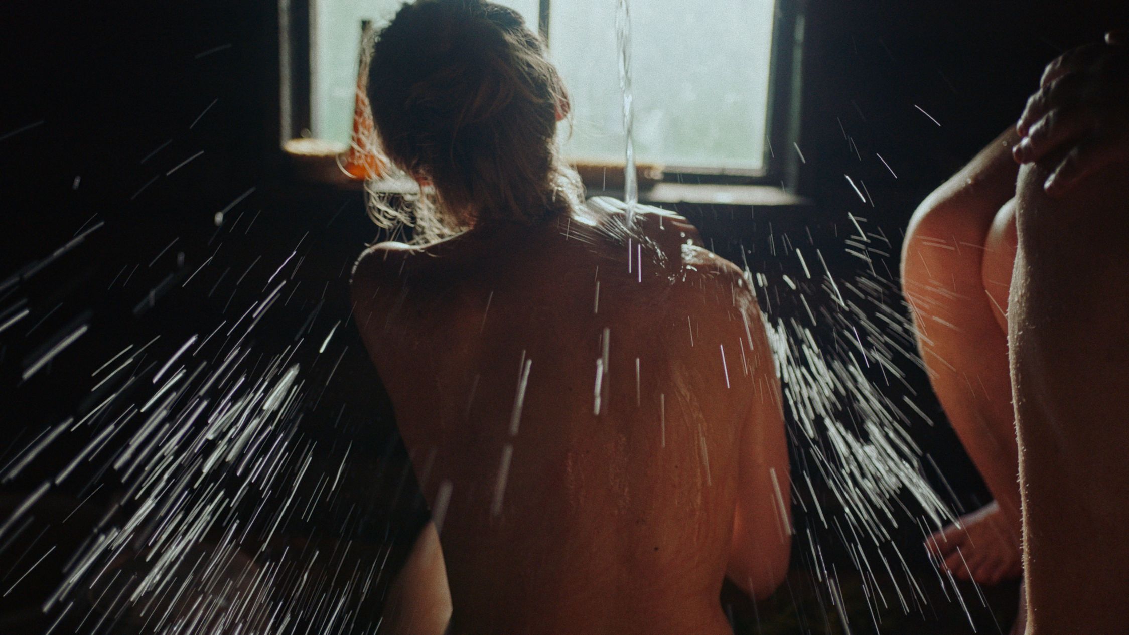 A close-up of a woman's back in a sauna, with water droplets cascading down their skin in a spray of light against a dark background. Sunlight filters through a window, highlighting the water's motion and creating a contrast with the shadows in the room. The focus on the water droplets gives the scene a dynamic and intimate quality.