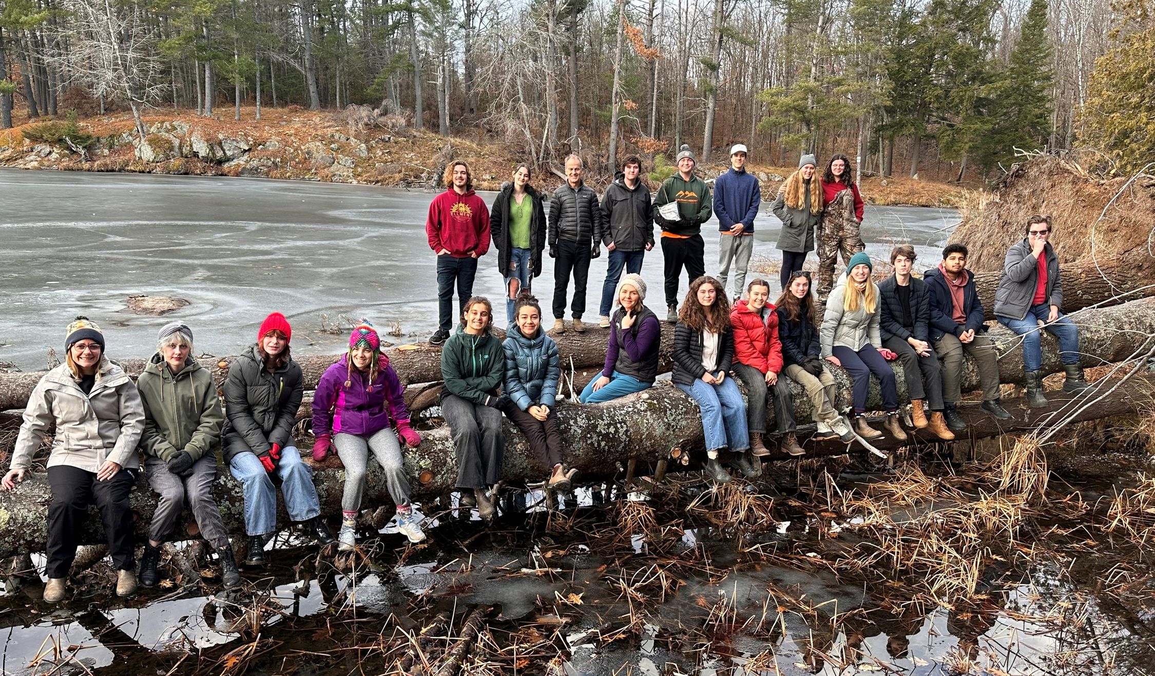 A group of twenty-two individuals of various attire are seated and standing on a fallen tree trunk over a body of frozen water, likely a lake or pond. The setting appears to be a cold day, as many individuals are wearing jackets, hats, and gloves. Bare trees and evergreens are visible in the background, suggesting a winter or late autumn season. The lighting suggests either early morning or late afternoon. There is no visible snow, but the ice on the water indicates freezing temperatures.