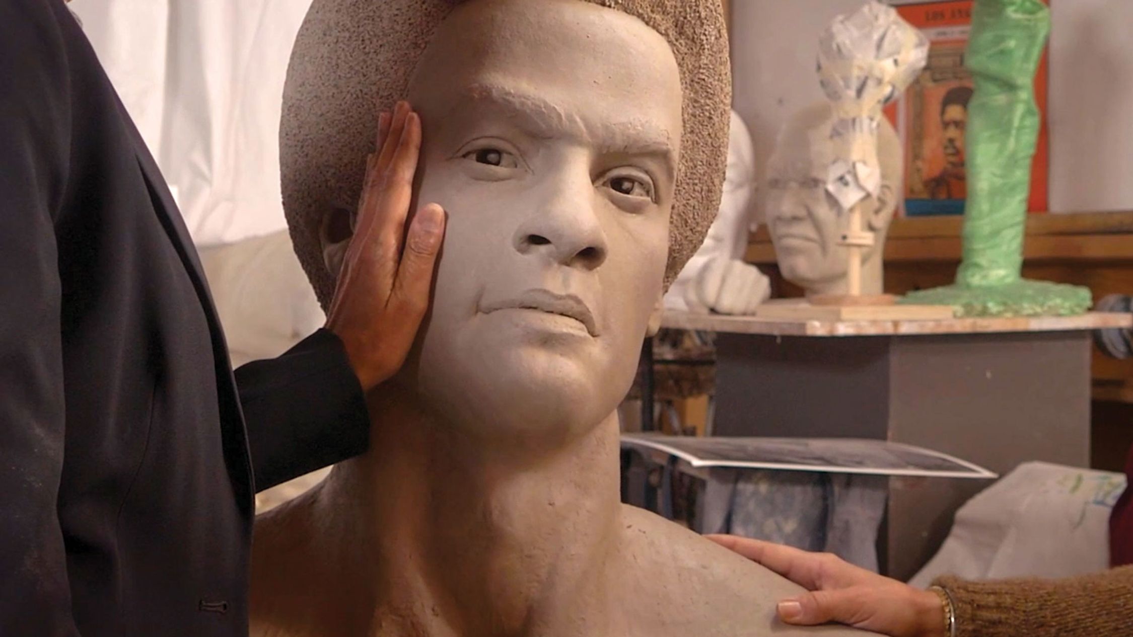 A person's hand is gently touching the cheek of a large, unfinished sculpted head and shoulders. The sculpture is highly detailed, with lifelike facial features, and it is mounted on a stand. In the background, there is an assortment of artistic materials and another bust sculpture, indicating that the setting is likely an artist's studio.