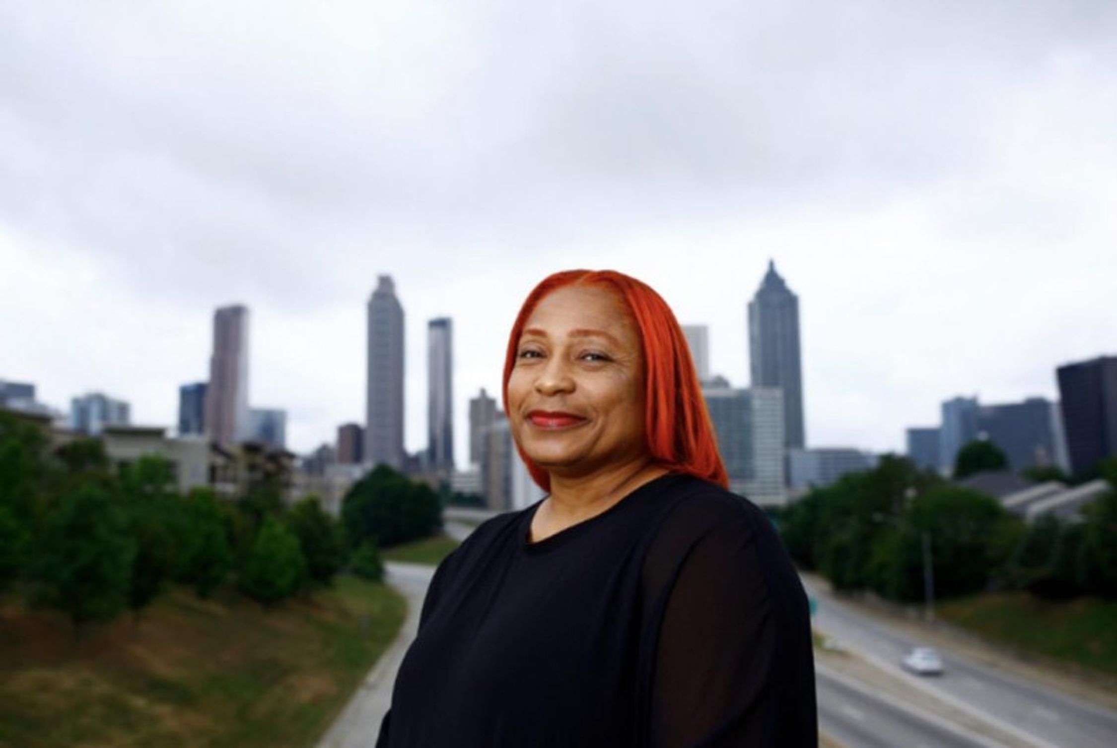 A person with shoulder-length bright red hair stands in the foreground, smiling slightly, with a blurred city skyline in the background. They are wearing a black top, and the overcast sky suggests a muted daylight setting. Below the skyline, there's a glimpse of a highway with moving vehicles, bordered by greenery. The focus on the individual with the cityscape backdrop creates a juxtaposition between the person and the urban environment.