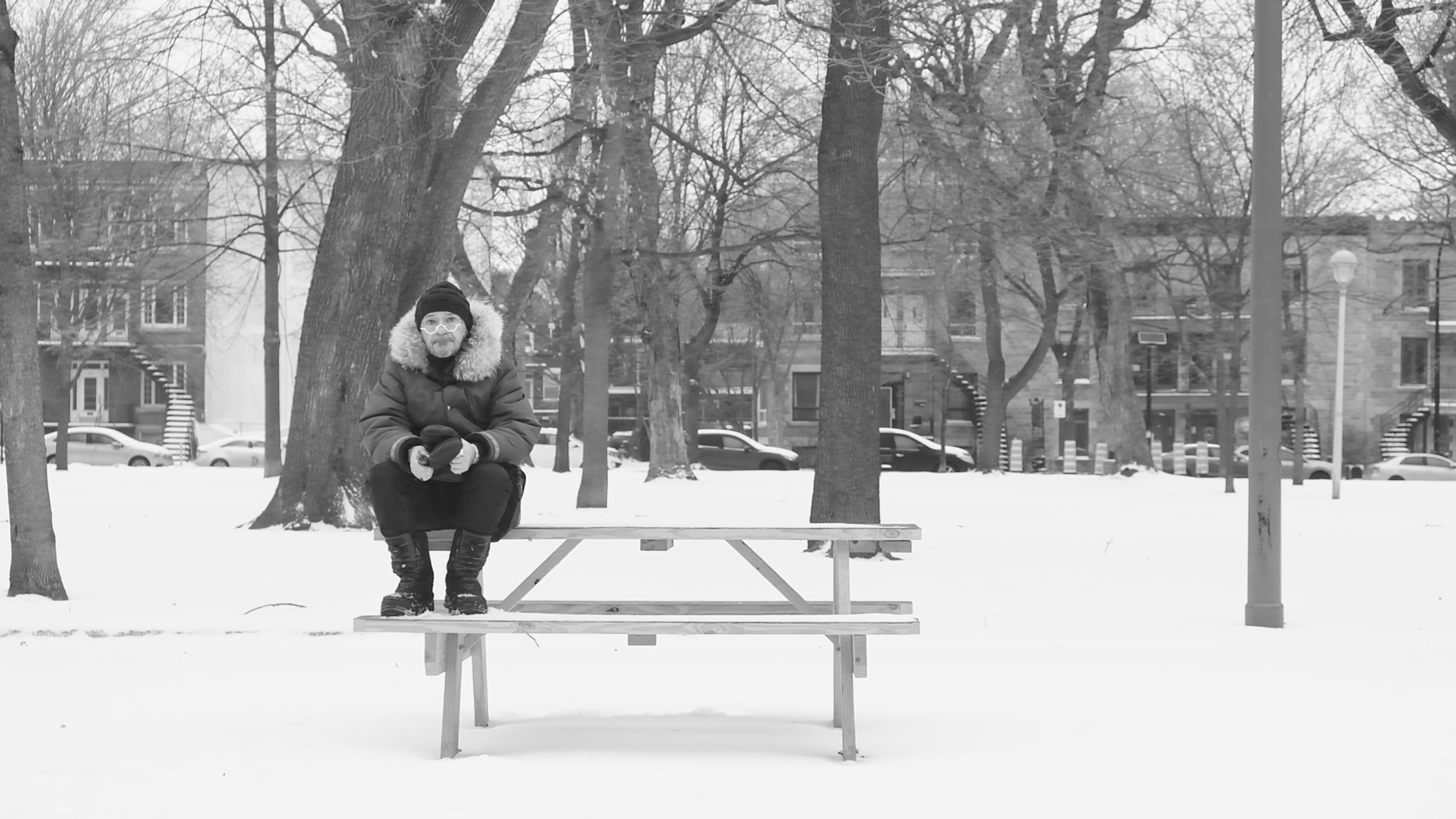 A black and white image of a snowy park setting, an individual is seated alone on a picnic table, dressed warmly in a heavy coat with a fur-lined hood, gloves, and a winter hat. The background features leafless trees, indicating it's a cold season, and buildings that suggest an urban environment. The person's posture and the empty park convey a sense of solitude or reflection.