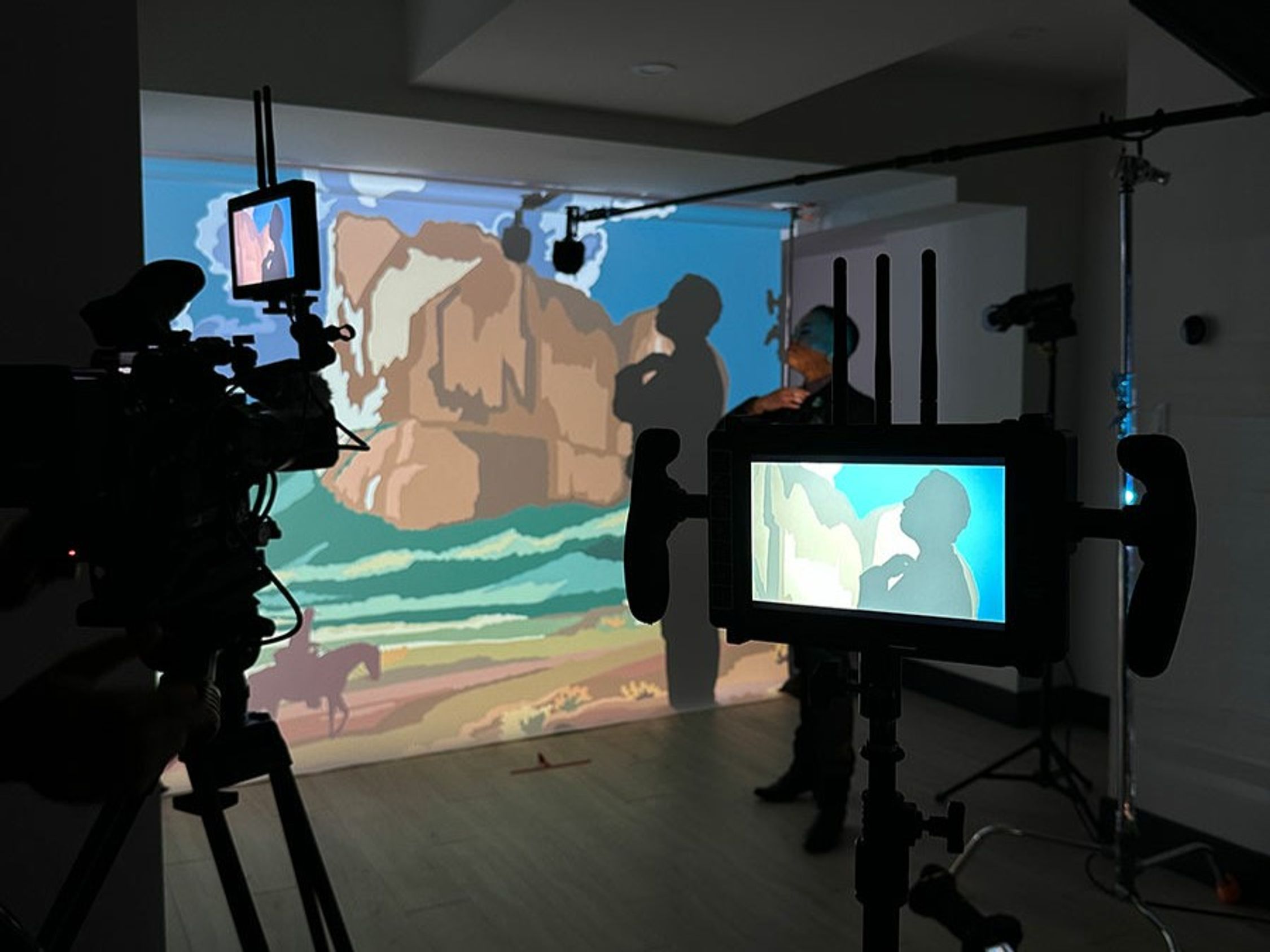 This image shows a behind-the-scenes view of a video production set. A video camera is in the foreground, focused on a person who is backlit against a large screen displaying colourful, stylized imagery resembling a landscape. The person's silhouette is also visible on the screen, indicating they might be performing or presenting. Monitors attached to the camera display the same image, allowing the crew to see what is being captured.