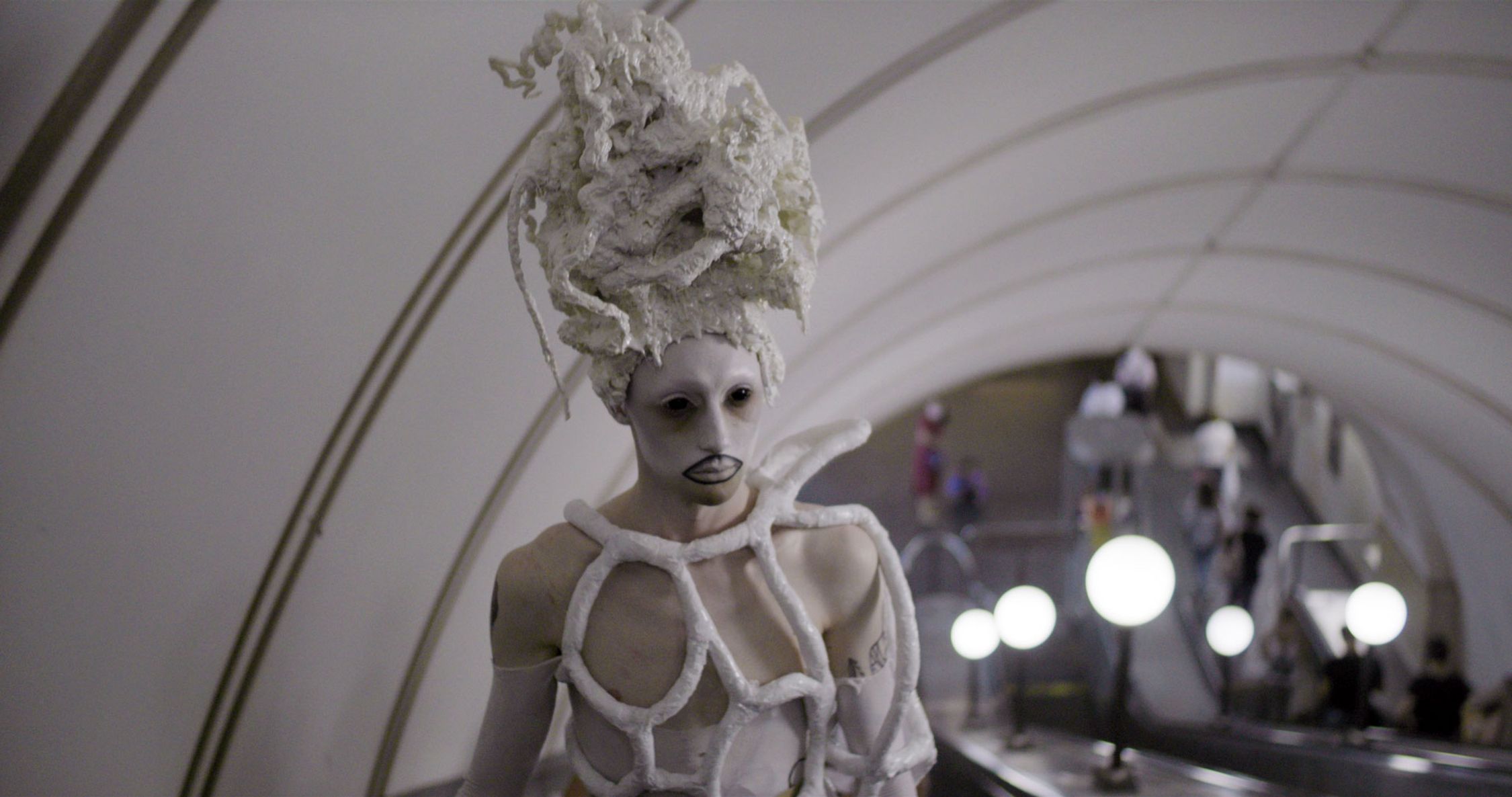 A figure with an avant-garde appearance is positioned in the foreground, dressed in what appears to be a white costume with structural, organic shapes enveloping the body and an elaborate headpiece resembling a stylized, textured wig or headdress. The figure's face is painted white, with darkened eyes and lips, and exhibits a neutral expression. The background shows an interior space with curved architecture and multiple people in motion, possibly a subway station or a public concourse, with a softly blurred focus that suggests movement and life happening around the still, striking figure in the foreground.