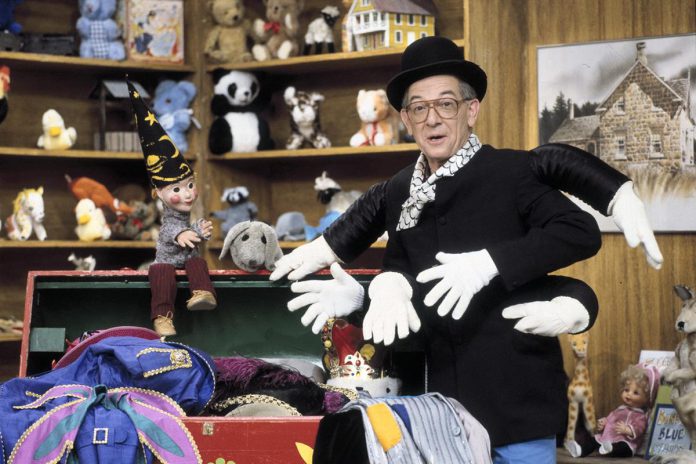 An image of a person wearing a top hat, glasses, and black and white scarf. To their left is a red tickle trunk with flowers painted onto it. The trunk is full of colourful clothes spilling over the edge. The person has on a fantastical black top, making it seem like they have six arms with white gloves covering the hands. In the background are shelves full of stuffed animals and other toys. 