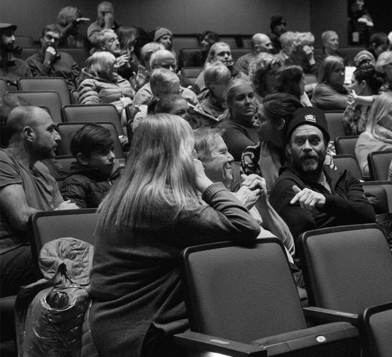 A black and white photo of ReFrame audience members talking to each other in a theatre. A few empty seats. Click here for more information about ReFrame Film Festival.