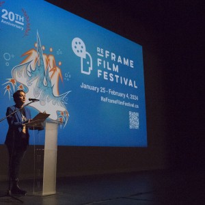 A person stands at a clear podium, speaking into a microphone. They are dressed in a blue jacket and dark pants, holding a clipboard. Behind them is a large projected screen displaying a bright blue background with the text “REFRAME FILM FESTIVAL” and event details: “January 25 - February 4, 2024” along with the website "ReFrameFilmFestival.ca" and a QR code. The screen also features a stylized abstract design with orange and blue accents, celebrating the festival's 20th anniversary, indicated by text and a laurel symbol in the top left corner.