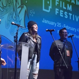 Three people stand on stage in front of microphones, smiling and speaking at the ReFrame Film Festival. From left to right: one person with long dark hair, wearing a black outfit with a blue and yellow sash; another wearing a black beanie, black leather jacket, and ripped light-wash jeans; and a third person with short dark hair, glasses, and a gray button-up shirt. Behind them is a large screen displaying the ReFrame Film Festival logo and event details, "January 25 - February 4, 2024," along with a QR code and abstract artwork in blue and green tones. The atmosphere is positive and engaging.