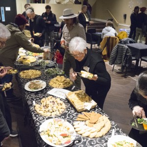 A group of people gathers around a long buffet table filled with a variety of finger foods, pastries, and small appetizers. The attendees are serving themselves from platters of food arranged on a black tablecloth with a swirling white pattern. Some are holding plates and chatting while others focus on selecting food. In the background, additional people are socializing in small groups, some seated at tables, while others stand near a staircase. Coats and jackets are draped over chairs, and the setting has a warm, casual atmosphere with dim lighting.