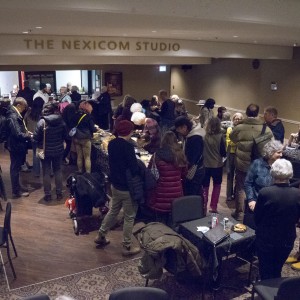 A large gathering of people fills a cozy studio space, labeled "THE NEXICOM STUDIO" in gold lettering above the entrance. Attendees are spread throughout the room, with many standing in groups near a long buffet table offering food, while others sit at tables, eating or reading. The atmosphere is social and relaxed, with people conversing, serving themselves, and mingling. Coats and jackets are draped over chairs, and a few people are seen wearing winter gear. The room features warm lighting, patterned rugs, and a mix of seated and standing guests, creating a bustling community event ambiance.