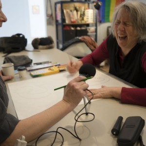 Two people are sitting at a table engaged in a lively, creative activity. The person on the right, with gray hair and wearing a red long-sleeve shirt under a dark vest, is laughing heartily. The person on the left, with shoulder-length blonde hair, is smiling and holding a microphone in one hand while drawing on paper with the other. Various art supplies, such as markers and pencils, are scattered on the table, along with cups and other personal items. In the background, shelves of books and a rainbow flag are visible, suggesting an informal, creative setting.