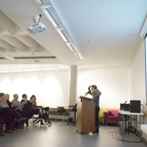 A speaker stands at a podium, addressing a seated audience in a well-lit room. The speaker is gesturing with raised arms, possibly emphasizing a point. The audience, consisting of several people, including some wearing face masks and one person with a walker, watches attentively, some applauding. A large projection screen to the right displays the "ReFrame Film Festival" logo and event details. The room is spacious, with a ceiling-mounted projector, and minimal furnishings like tables with audio equipment and a colorful couch in the background. The setting appears to be part of a film festival presentation or talk.