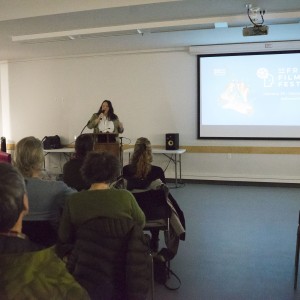 A speaker stands at a podium in front of an audience, delivering a presentation. The room is dimly lit, and a large screen to the speaker's right displays the "ReFrame Film Festival" logo, along with the event dates (January 25 – February 4, 2024) and a QR code. The audience is seated in rows, facing the speaker, with people of varying ages. The speaker, holding a microphone, is mid-speech, gesturing with one hand. The room is simple, with tables along the walls holding audio equipment, bags, and other personal items. A pink couch is visible in the background.