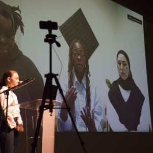 A person stands at a clear podium, speaking into a microphone while facing an audience. In the background, a large video call is projected onto a screen, showing three individuals participating remotely. One person, with long braids and glasses, is speaking with expressive hand gestures. Another individual, wearing a hijab, sits attentively beside them, while a third person appears partially visible on the left, looking down. A tripod with a camera is positioned in the foreground, capturing the scene. The setup suggests a hybrid event, blending in-person and virtual participants, likely for a presentation or panel discussion.