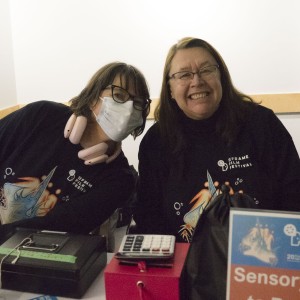 Two individuals sit at a table, smiling and wearing matching "ReFrame Film Festival" shirts. One person on the left is wearing a face mask and pink headphones around their neck, while the other person on the right smiles widely without a mask. In front of them are several items, including a cash box, a large calculator, and a small red box. A sign in the foreground reads "Sensory Kits for Rent." The setting suggests they are at a festival booth or information desk, engaged in welcoming or assisting attendees. The lighting is soft, with a friendly and approachable atmosphere.