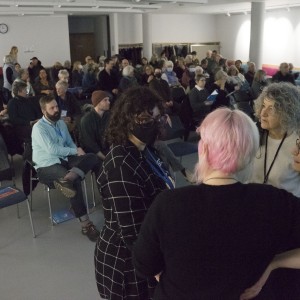 A large group of people is seated in a well-lit room, awaiting the start of an event. In the foreground, four individuals stand in a small group, engaged in conversation. One person has pink hair, another wears a black mask, and the others have curly and short gray hair, respectively. The audience behind them consists of a mix of older and younger attendees, some wearing masks. A few people are still finding seats, while others are chatting. Empty seats with "Reserved" signs are scattered throughout the room. The atmosphere feels anticipatory, with people settling in for a presentation or talk.