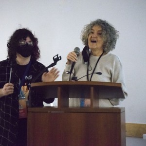 Two people stand at a podium, one speaking into a microphone. The person on the right, with curly gray hair, is smiling and speaking while holding the microphone, with a black face mask hanging around their neck. The person on the left, with dark curly hair and glasses, is wearing a face mask and holding a second microphone, seemingly assisting or preparing to speak. Both are wearing lanyards, and the person on the left’s lanyard reads "STAFF." The setting appears casual, with a collaborative and friendly atmosphere as the two prepare or conduct a presentation.