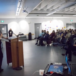 A speaker stands at a podium, holding a microphone, addressing an audience seated in a room with a projector screen. The speaker is facing the audience, and a blue presentation slide on the screen reads "ReFrame Film Festival" with additional text and graphics. The audience, composed of mostly older adults, is attentively watching, some wearing masks. The room is well-lit, with white walls and ceiling-mounted lights. There are tables in the background with various items, including a laptop and audio equipment. A person in dark clothing stands near the back of the room, observing the scene.