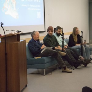 A panel of four individuals sits on a light blue couch, participating in a discussion during an event. Each person holds or is seated near a microphone. The person on the left is speaking, reading from papers in their hand, while the others listen. One panelist wears a red beanie, while the others are dressed casually. A moderator, standing at a wooden podium, looks towards the panel, overseeing the discussion. In the background, a large projection displays "ReFrame Film Festival" details on the screen. The setting suggests a casual, interactive panel discussion as part of the festival.