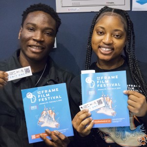 Two smiling individuals hold up tickets and programs for the "ReFrame Film Festival." Both are holding blue festival booklets featuring the event's name, logo, and dates (January 25 – February 4, 2024). The person on the left is holding a ticket that says "Admit One" alongside their program, while the person on the right holds both items similarly. They are standing in front of a dark blue wall, with some posters partially visible in the background. Both appear excited and happy, showcasing their involvement in the event.