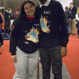 Two individuals stand side by side, smiling and posing for the camera. Both are wearing matching "ReFrame Film Festival" long-sleeve shirts with colorful designs on the front. The person on the left wears gray sweatpants and black boots, while the person on the right wears camouflage-patterned pants and glasses. They have their arms around each other, appearing happy and relaxed. The background features a red carpeted floor, with other festival attendees visible behind them, engaged in various activities in a social setting. The atmosphere is lively and friendly.