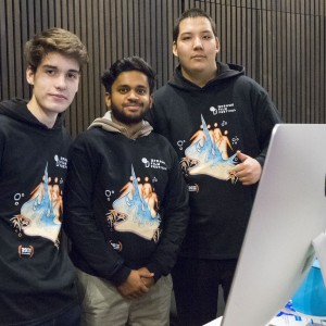 Three individuals stand behind a table, posing together for a photo. All are wearing matching black "ReFrame Film Festival" hoodies with colorful designs on the front. They appear to be festival volunteers or attendees. One of them is standing next to a large computer monitor, and the table in front of them holds various items, including bags with blue ribbons and festival materials. The background consists of dark, vertical wood paneling, giving a professional setting to the space. The group looks engaged.