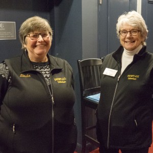 Two older women are standing indoors, smiling at the camera. Both are wearing black vests with "Showplace Volunteer" embroidered in yellow. The woman on the left has short grayish-brown hair, glasses, and a name tag that reads "Linda." She is wearing a black and white patterned sweater under her vest. The woman on the right has short white hair, glasses, and a white turtleneck beneath her vest. She has a name tag reading "Audrey." Behind them, there are tall black chairs and a dark-colored wall. The floor is covered with red carpeting.