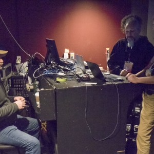 Three individuals are gathered around a control desk in a dimly lit room, likely operating audio-visual or technical equipment. The one on the left is seated, wearing a brown cap, gray jacket, and jeans. The two on the right are standing; one has a gray beard and curly hair, wearing a black hoodie, while the other has short hair, wearing a black shirt and tan pants with a mask pulled down around his chin. The desk is cluttered with various laptops, tangled cables, and technical gear. The wall behind them is a reddish color, and there are coffee cups and water bottles on the desk.