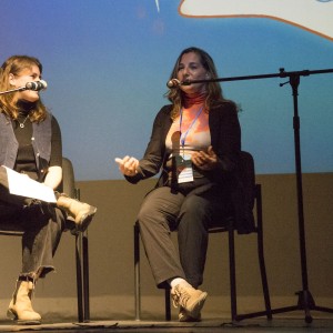 Two women are seated on a stage, each in front of a microphone, engaged in a conversation. The woman on the left has shoulder-length hair, is wearing a black top with a blue vest, ripped black jeans, and tan boots. She is holding a paper on her lap. The woman on the right is speaking, gesturing with her hands. She has long hair and is wearing a black blazer over a patterned orange and white top, black pants, and tan shoes. Both women are seated on simple chairs. A large blue projection with abstract shapes is visible in the background.