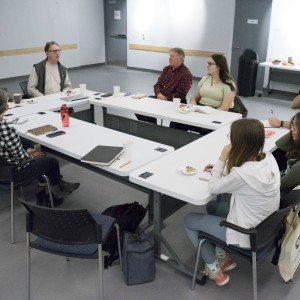 In this overhead view of a group meeting, ten participants are seated around a U-shaped table setup in a well-lit room. The group is engaged in discussion, with one person in the center speaking while others listen or take notes. Participants are casually dressed, with notebooks, water bottles, and coffee cups on the tables. The person speaking appears to be leading the conversation, while others focus on the discussion or jot down notes. A table with snacks and drinks is visible against the wall in the background. The room has a clean, minimalist feel, with a professional yet informal atmosphere.