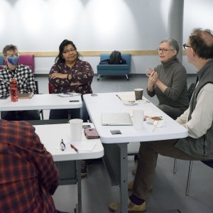 Five individuals are seated around a table in a casual meeting setting. The person on the right, wearing a gray turtleneck, is speaking and gesturing with their hands, while others listen attentively. One person, second from the left, is wearing a patterned shirt with arms crossed, and another individual, next to them, is wearing a checkered shirt with a blue face mask. The table is scattered with coffee cups, notebooks, and a water bottle, creating a relaxed yet focused environment. In the background, colorful lounge chairs and a backpack are visible.