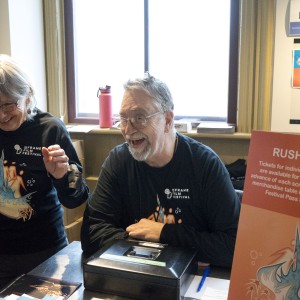 Two volunteers, both wearing black "ReFrame Film Festival" shirts with colorful graphics, stand behind a ticket booth, smiling and interacting warmly. The person on the right. wearing glasses and a gray beard, leans forward, appearing to laugh or engage in conversation. In front of him is a cash box and festival materials. The person on the left, with short gray hair and glasses, is also smiling and gesturing with her hands. A sign next to them reads "Rush Tickets" with information about ticket pricing and availability. The setting has a friendly, welcoming atmosphere, and a bright window behind them illuminates the booth.