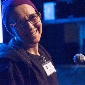 A person is smiling warmly at the camera while standing near a microphone. They are wearing glasses, a purple beanie, and a dark hoodie. A name tag is visible on their chest, clipped to the hoodie, though the text is slightly blurred. The lighting in the scene is dim, with a soft blue hue illuminating the background, in a stage/event setting. A bright screen is partially visible in the left background. The overall mood appears relaxed and casual.