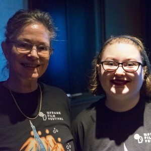 Two individuals, both wearing "ReFrame Film Festival" t-shirts, smile for the camera in a dimly lit setting. The person on the left has glasses and graying hair tied back, wearing a necklace. The person on the right, also wearing glasses, has short, light brown hair and is smiling brightly. The lighting is low but casts a warm glow on their faces, with a blue background adding to the ambiance.